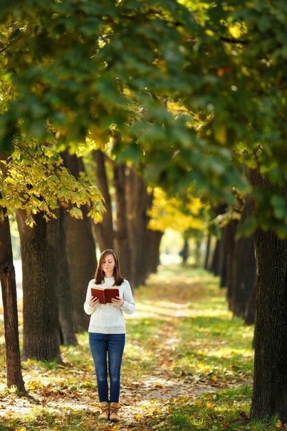 La hermosa mujer de pelo castaño sonriente feliz en suéter blanco de pie con un libro rojo en el parque de la ciudad de otoño en un día cálido. Otoño de hojas doradas. Concepto de lectura.