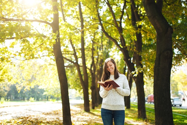 La hermosa mujer de pelo castaño sonriente feliz en suéter blanco de pie con un libro rojo en el parque de la ciudad de otoño en un día cálido. Otoño de hojas doradas. Concepto de lectura.