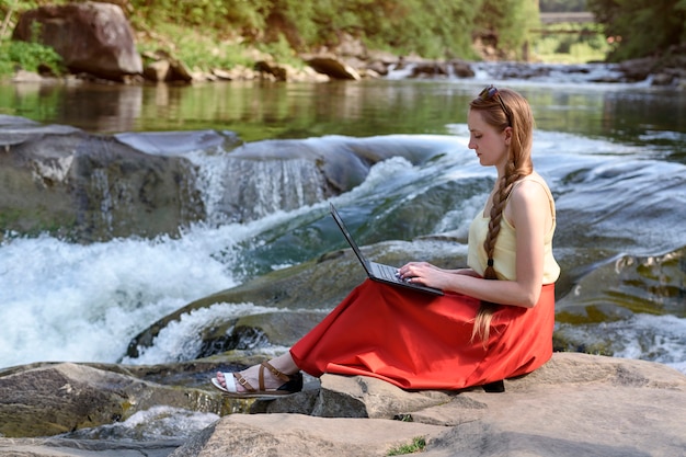 Hermosa mujer de pelo largo en falda roja con portátil sentado en una roca, cascada del río de montaña. Concepto independiente. Trabajo en la naturaleza