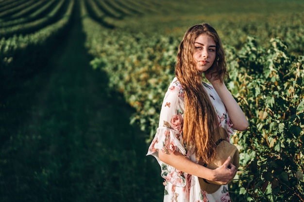 Hermosa mujer con pelo largo y rizado y cara de pecas en campo de grosella. Chica con un vestido ligero camina en el día soleado de verano