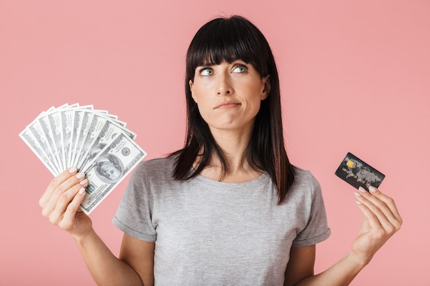 una hermosa mujer de pensamiento increíble posando aislada sobre la pared de la pared de color rosa claro con dinero y tarjeta de crédito.