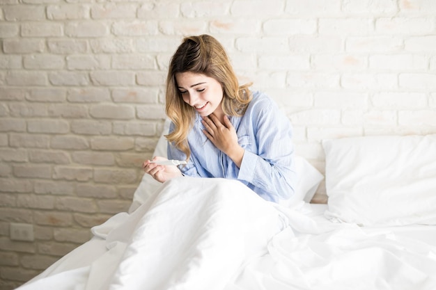 Foto hermosa mujer en pijama sentada en la cama y mirando una prueba de embarazo positiva y sintiéndose emocionada