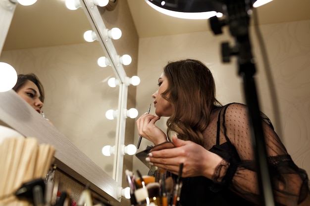 Una hermosa mujer pinta sus labios de cerca mirando en un espejo con lámparas, primer plano.