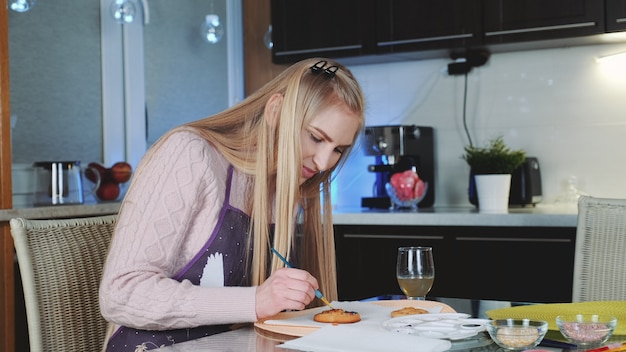 Hermosa mujer pintando galletas dulces con colorantes alimentarios especiales en la cocina de su casa