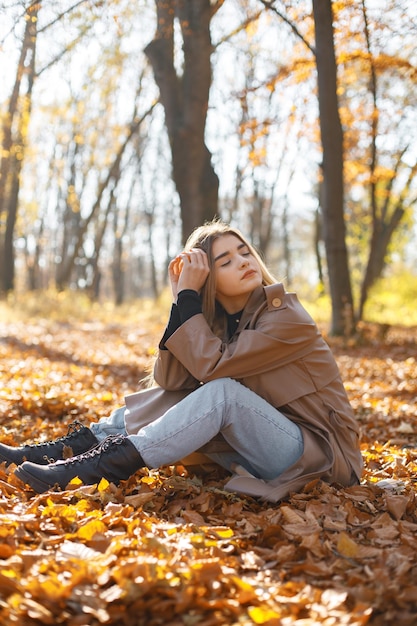 Hermosa mujer posando para una foto en el parque de otoño. Niña sentada sobre hojas amarillas. Mujer rubia con abrigo beige.