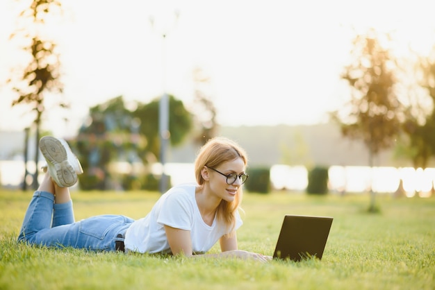 Hermosa mujer que trabaja con la computadora portátil en el parque sobre la hierba. El concepto de trabajo a distancia