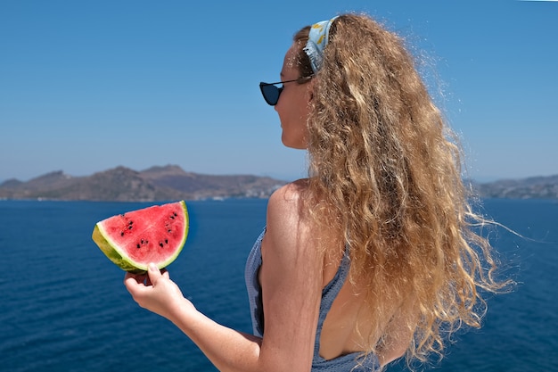 Hermosa mujer con una rebanada de sandía con traje de baño comiendo una rebanada de sandía madura roja con vistas al mar en verano.