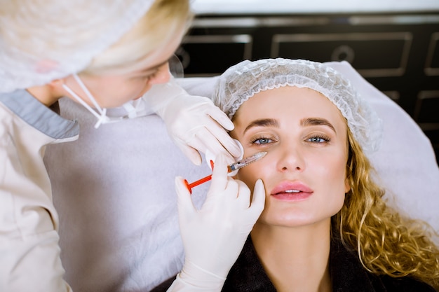 Hermosa mujer recibe inyecciones. Cosmetología. Cara de belleza. Ciérrese encima del retrato de la mujer rubia con las manos femeninas de la cosmetóloga cerca de su cara