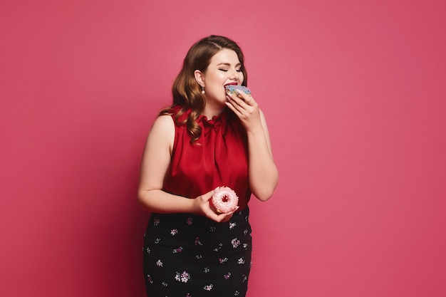 Hermosa mujer regordeta con maquillaje brillante en traje de moda comiendo deliciosas donas sobre fondo rosa es ...