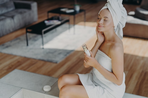 Hermosa mujer relajada envuelta en una toalla aplicando crema facial