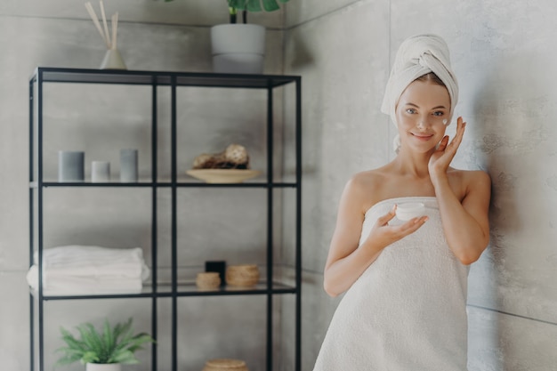 Hermosa mujer relajada envuelta en una toalla aplicando crema facial