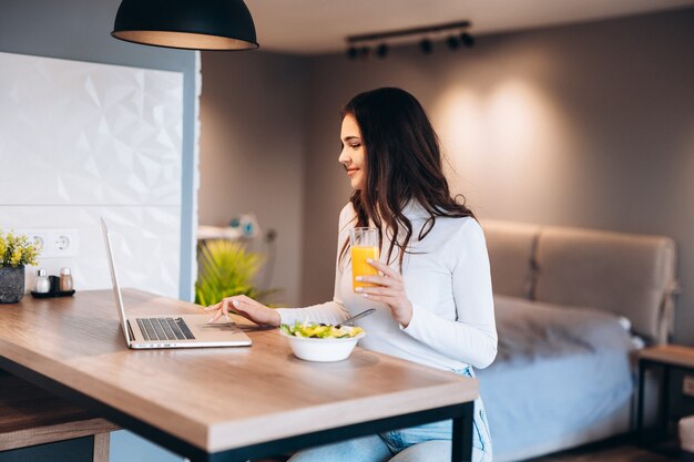 Hermosa mujer relajante con su computadora portátil mientras sostiene un vaso de jugo de naranja en la cocina. Trabajando desde casa en cuarentena. Distanciamiento social Autoaislamiento.