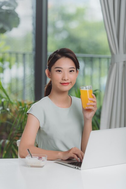 Hermosa mujer de relax con su computadora portátil mientras sostiene un vaso de jugo de naranja