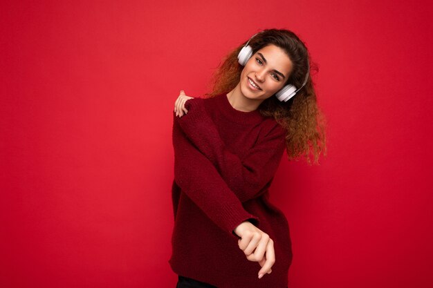 Hermosa mujer rizada morena joven sonriente feliz con suéter rojo oscuro aislado sobre la pared de fondo rojo con auriculares bluetooth blancos escuchando música y divirtiéndose mirando a la cámara