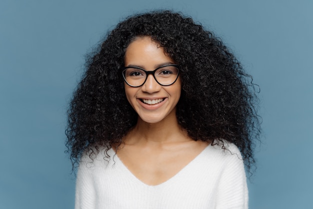 Hermosa mujer rizada con una sonrisa agradable, usa gafas y un jersey informal blanco