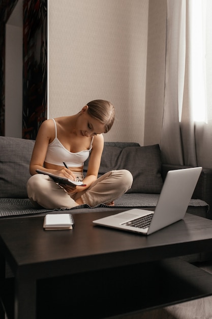 Hermosa mujer en ropa casual está sentada en un sofá en un apartamento y se dedica al autoaprendizaje, toma notas en un cuaderno junto a una computadora