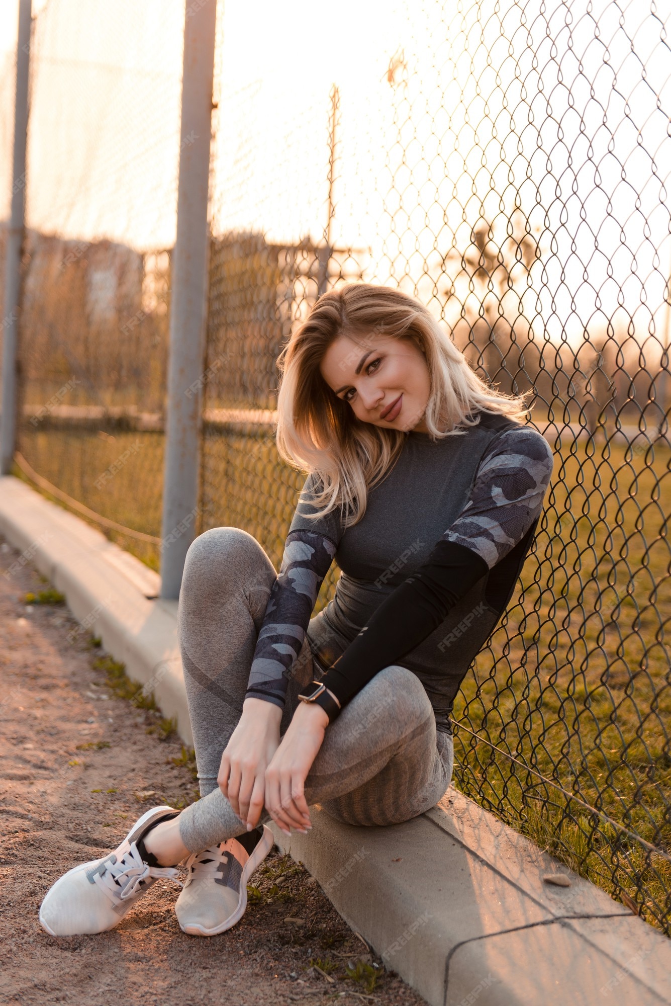 Hermosa mujer en ropa deportiva gris ajustada sonriendo y mirando a la cámara. deportiva mujer rubia sentada acera cerca de la valla de concepto estilo de vida saludable.