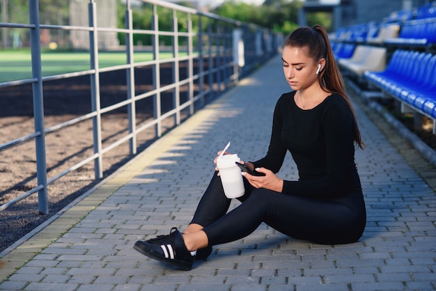 Una hermosa mujer en ropa deportiva negra bebe nutrición deportiva de una botella blanca y usa el teléfono en la tribuna del estadio después del entrenamiento. Concepto deportivo y saludable.