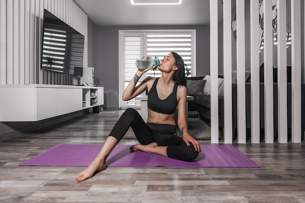 Hermosa mujer en ropa deportiva se sienta en una estera de yoga y bebe agua