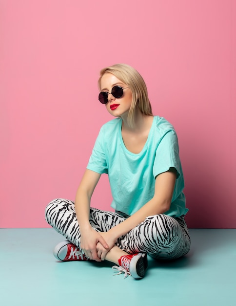 Hermosa mujer en ropa de estilo y gafas de sol sentado en el piso azul cerca de la pared rosa