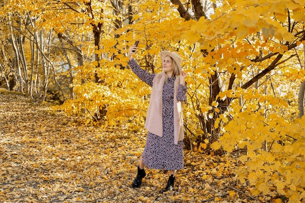 Hermosa mujer en ropa de moda y sombrero toma selfie en teléfono cerca del árbol de otoño amarillo