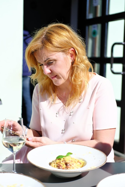 Hermosa mujer rubia adulta se sienta en una mesa en un café en la calle y mira la pantalla de un teléfono móvil con una mirada seria Foto vertical