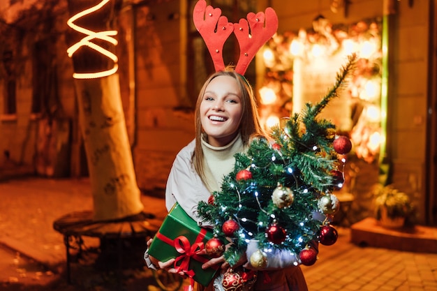 Hermosa mujer rubia con un árbol de Navidad y un regalo en sus manos sonriendo