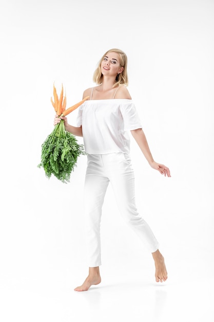 Hermosa mujer rubia con una blusa blanca sosteniendo una zanahoria fresca con hojas verdes sobre un fondo blanco. Salud y Dieta