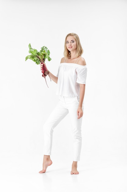 Hermosa mujer rubia con una blusa blanca sostiene una remolacha con hojas verdes sobre un fondo blanco. Salud y vitaminas