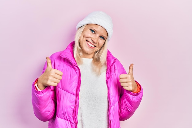 Hermosa mujer rubia caucásica con sombrero de lana y signo de éxito de abrigo de invierno haciendo un gesto positivo con la mano, pulgares arriba sonriendo y feliz. expresión alegre y gesto ganador.