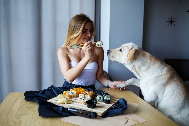 Hermosa mujer rubia comiendo primer plano de sushi con su perro labrador, mujer sonriente con maquillaje encantador sosteniendo un rollo de sushi con palillos. Comida japonesa saludable. Concepto de dieta