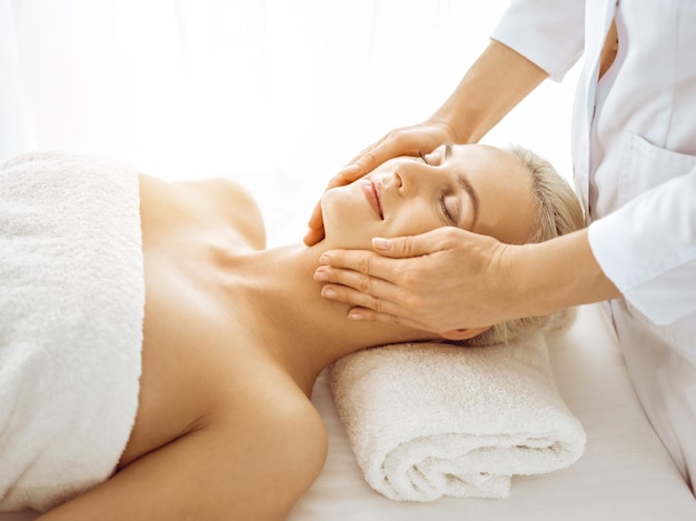 Una hermosa mujer rubia disfrutando de un masaje facial con los ojos cerrados en un centro de spa soleado. Tratamiento relajante en conceptos de medicina y belleza.