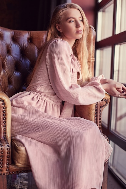 Hermosa mujer rubia elegante en vestido rosa sentado en un sillón de cuero