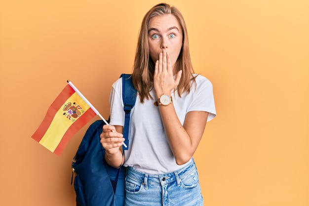 Hermosa mujer rubia estudiante de intercambio con bandera española cubriendo la boca con la mano sorprendida y asustada por error expresión sorprendida