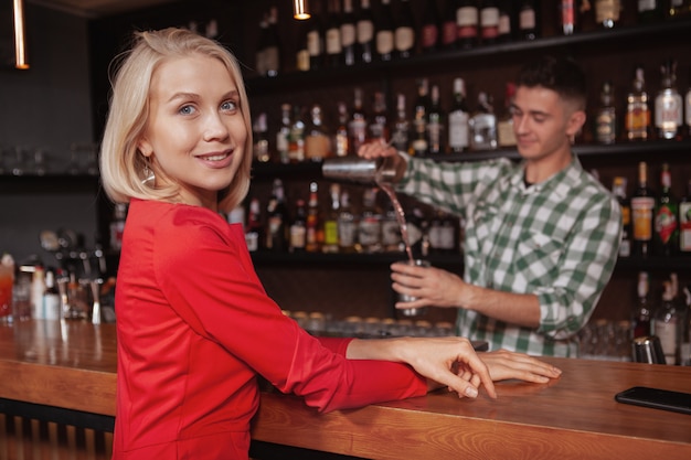 Hermosa mujer rubia feliz sonriendo a la cámara, esperando su cóctel en el bar