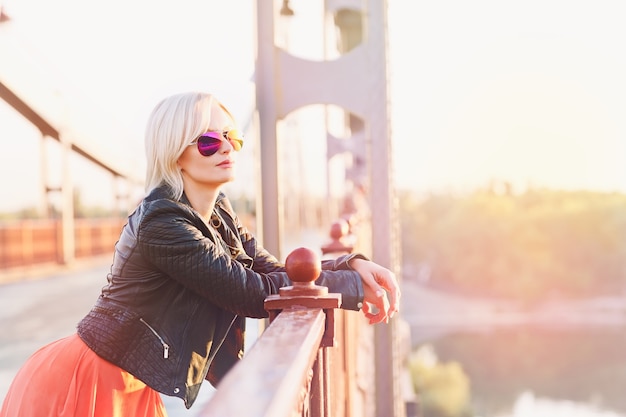 Hermosa mujer rubia en gafas de sol rosas posando en el puente al atardecer