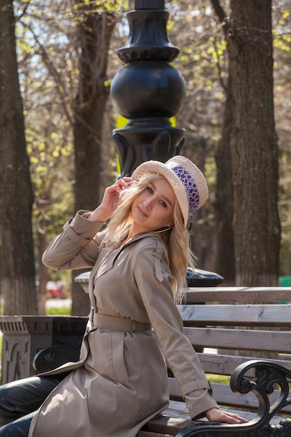 Hermosa mujer rubia con impermeable y sombrero se sienta en un banco