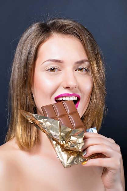Hermosa mujer rubia joven desnuda con maquillaje brillante comiendo barra de chocolate.