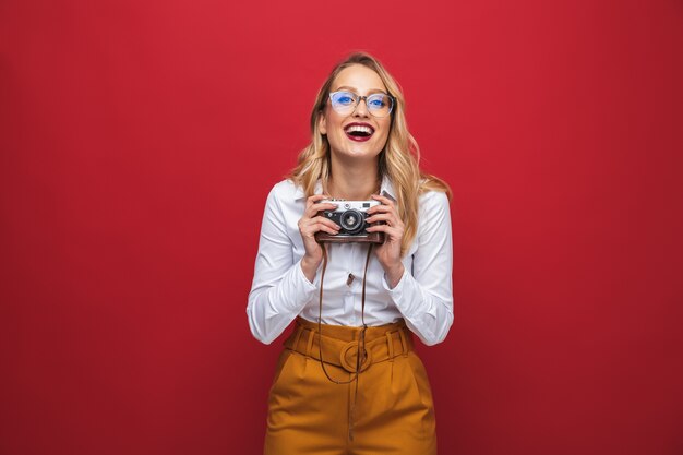 Hermosa mujer rubia joven feliz que se encuentran aisladas sobre fondo rojo, sosteniendo la cámara de fotos