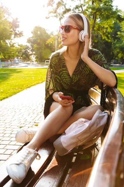 Hermosa mujer rubia joven feliz sentada en un banco en el parque de la ciudad, escuchando música con auriculares inalámbricos