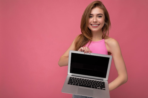 Foto hermosa mujer rubia joven sonriente sosteniendo el ordenador portátil con pantalla de monitor vacía vistiendo rosa crop top