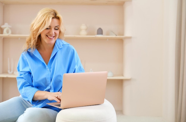 Hermosa mujer rubia madura con una camisa azul sentada en un sofá con una laptop en un interior de color beige mira la pantalla y sonríe