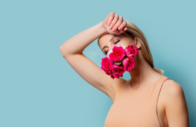 Hermosa mujer rubia en mascarilla con rosas sobre fondo azul.