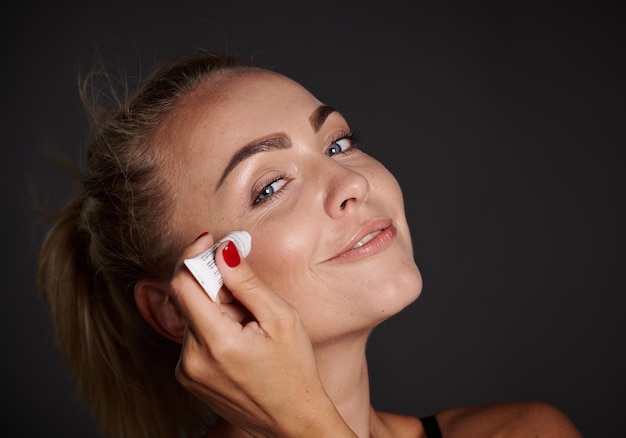 Hermosa mujer rubia de mediana edad exprimiendo un poco de crema suavizante antiarrugas debajo de los ojos para evitar la hinchazón y prevenir el proceso de envejecimiento temprano Antienvejecimiento spa rejuvenecedor belleza cuidado de la piel juvenil