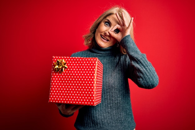 Hermosa mujer rubia de mediana edad sosteniendo un regalo de cumpleaños sobre un fondo rojo aislado con cara feliz sonriendo haciendo el signo de ok con la mano mirando a través de los dedos