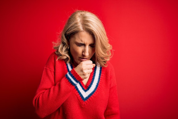 Hermosa mujer rubia de mediana edad con suéter casual sobre fondo rojo aislado sintiéndose mal y tosiendo como síntoma de resfriado o bronquitis Concepto de atención médica