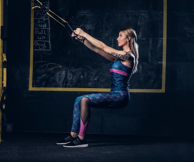 Hermosa mujer rubia musculosa en ropa deportiva con un tatuaje en el brazo haciendo ejercicio con el sistema TRX en el gimnasio. Concepto TRX.