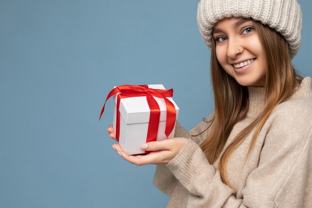 Hermosa mujer rubia oscura joven feliz aislada sobre pared colorida vistiendo ropa casual elegante con caja de regalo y mirando a la cámara