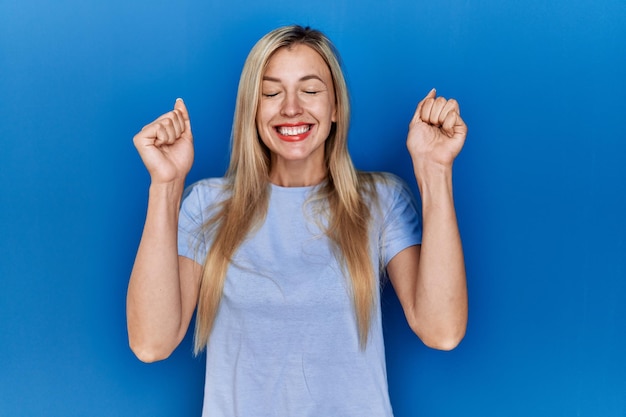 Hermosa mujer rubia con pantalones casual sobre fondo azul emocionada por el éxito con los brazos levantados y los ojos cerrados celebrando la victoria sonriendo. concepto de ganador.