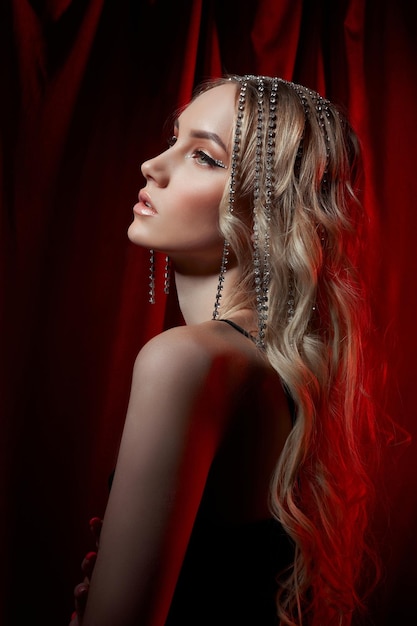 Hermosa mujer rubia con el pelo largo sobre un fondo de terciopelo rojo. Joyas para el cabello en la cabeza de una mujer, maquillaje moderno y flechas en los ojos.